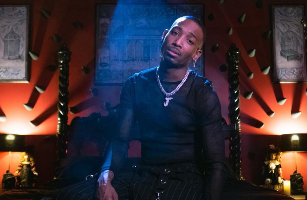 Promotional photo for "All I Need Forever" which sees J Young MDK sitting on a stage with low light in the room, wearing a black t-shirt and pants, with a silver necklace.
