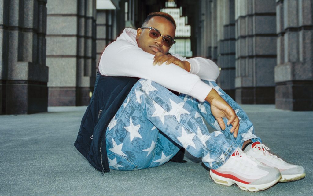 Promotional photo for "555" which sees Jay Americana sitting on a concrete floor with his arms around his knees. He is wearing jeans with white stars on, some white Nikes, a black jacket with white sleeves and some sunglasses.