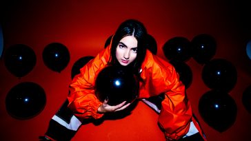 Promotional photo for "Dead Body" which sees KEHLI wearing an orange baggy jumpsuit, sitting on a red floor, looking up at the camera whilst holding a black balloon close to her chest. There are also various black balloons around her too.