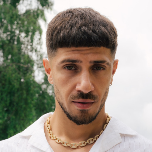 Artist profile image of Marcello Spooks who has just released "Endless Drive: A Love Story" EP. He is earing a white polo shirt with a gold chain necklace. He's got a stubble beard and moustache, and his hair is short. There's green trees behind him.