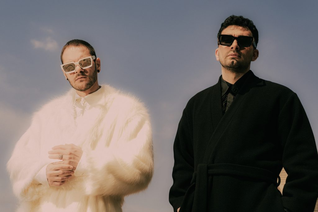 Promotional photo for "Artificial Intelligence" which sees a professional photo studio shot of Marnik posing. The left guy is wearing a white jumper, and the right guy is wearing a black wool overcoat. They are both wearing shades with the one on the left wearing pink shades and the one on the right wearing black shades. The background is grey.