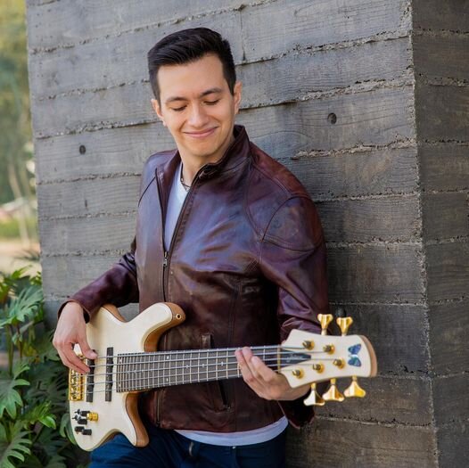 Photo for "Tower In The Sky" which sees Mauricio Morales wearing a brown leather jacket with his back against a grey brick wall, playing a bass guitar.