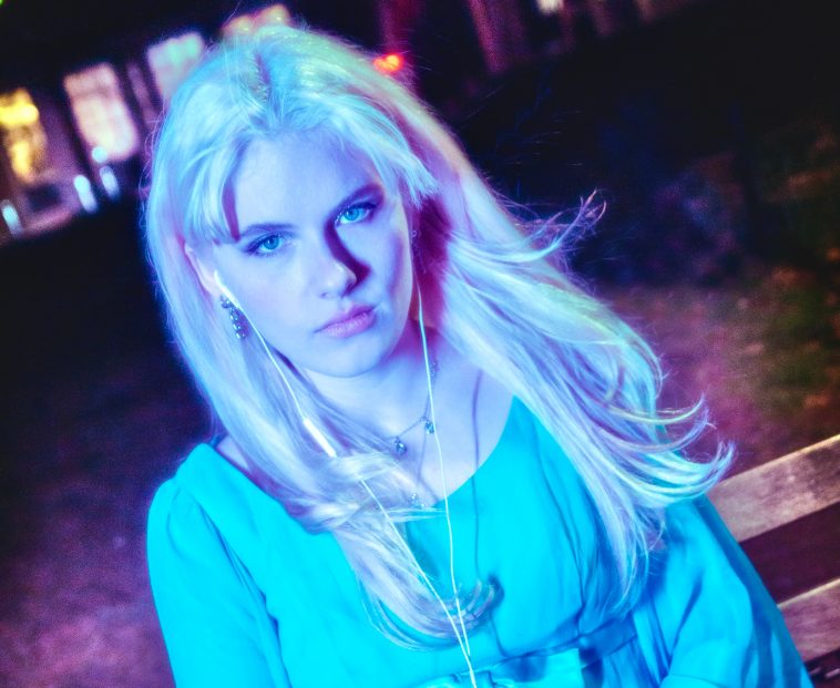 Promotional photo for "breakup with a friend" which sees Meg Smith wearing a baby-blue dress, sitting on a bench looking at the camera as the wind blows her mid-length blonde hair. She is sitting in a park with trees and leaves behind her, and the photo is slightly edited to create a glowing effect around Meg Smith.