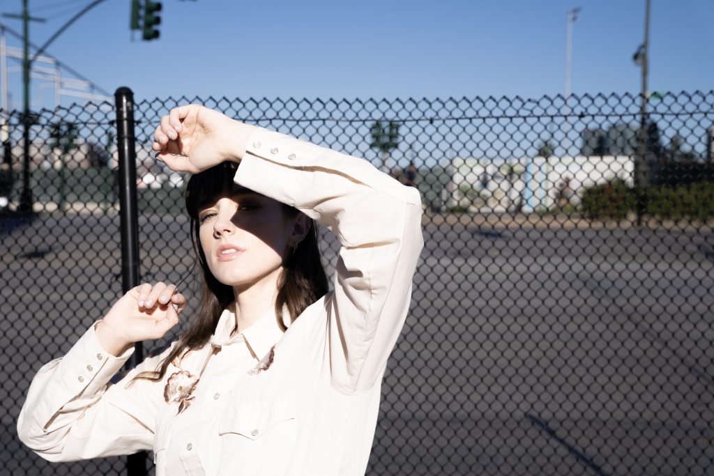 Promotional photo for "Wounds (Healing)" which sees Olivia Reid with a chained fence behind her, shielding her eyes from the sun with her left arm. She is wearing a light pink shirt.