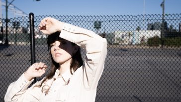 Promotional photo for "Wounds (Healing)" which sees Olivia Reid with a chained fence behind her, shielding her eyes from the sun with her left arm. She is wearing a light pink shirt.