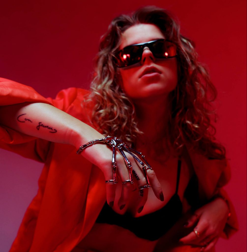 Promotional photo for "kinda like it" which sees REYA with dirty blonde wavy hair that falls past her shoulders and huge sunglasses. She's wearing a red jacket over a black bra and has a intricate hand bracelet that is attached onto the back of her right hand. She's got her right arm up shoulder-height, with her hand extended out in front of her.