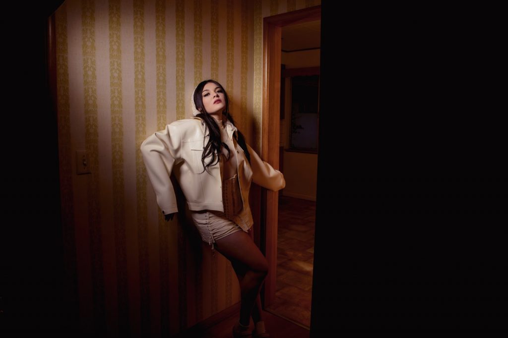 Promotional photo for "Hooked" which sees Sam Short in a cream short dress and an oversize white mac jacket, leaning against a striped wallpaper wall, with an open doorway to the right of her. The room is dimly lit and and brown hair cascades down past her shoulders.