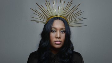 Promotional photo for "Black British" which sees V V Brown looking at the camera with her black hair flowing past her shoulders and past her black jumper. She is wearing a huge gold spikey headdress crown and the background is a plain grey.