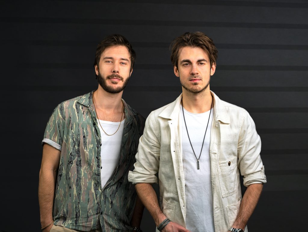 Promotional photo for "Dopamine Junkie" featuring Ben Samama, which shows the DJ duo Vicetone standing against a dark wood wall, with their hands in their pockets. They are both wearing shirts, the one on the left is wearing a sea-green washed shirt, opened half-way with a white vest underneath, and the guy on the right is wearing a cream shirt over a white tee with a dark metal necklace.