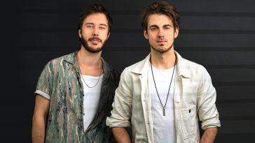 Promotional photo for "Dopamine Junkie" featuring Ben Samama, which shows the DJ duo Vicetone standing against a dark wood wall, with their hands in their pockets. They are both wearing shirts, the one on the left is wearing a sea-green washed shirt, opened half-way with a white vest underneath, and the guy on the right is wearing a cream shirt over a white tee with a dark metal necklace.