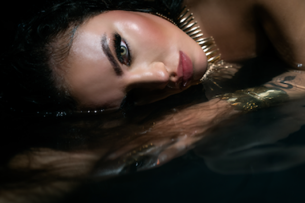 Promotional photo for "You Already Know" which sees AKICITA lying in some water. It's a close-up of her face which is just above the water's surface, her black hair is behind her, and her make-up is on-point. She's wearing a gold lined choker necklace and she has her hand just in front of her face, but under the water's surface.