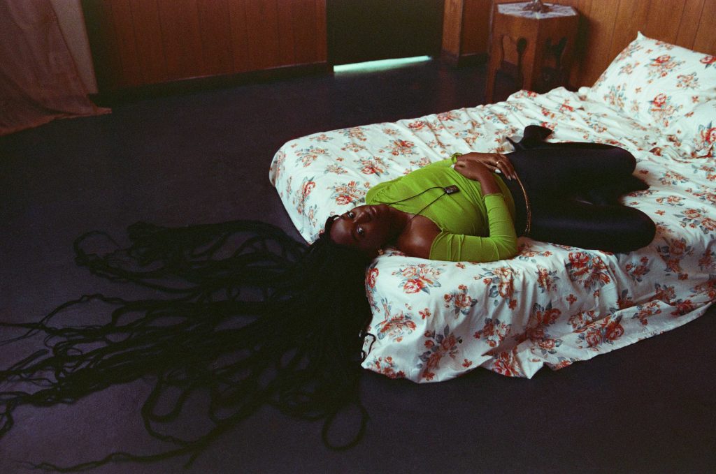 Promotional photo for "To It" which sees Aryeè The Gem lying on her back on a white bed with her massively long braids spread out on the floor at the end of the bed, which is where her head is. She's wearing a green tube top and black leggings, with a gold chain with a pendant on the end, resting over her heart.