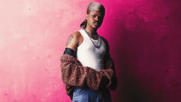 Promotional photo for "I Salute" featuring Zlatan, which sees Bad Boy Timz standing in front of a pink background, wearing a white vest under a silver chain, with his brown jacket undone and off his shoulders, with his arms still in the sleeves. He's also wearing blue jeans, and a gold and black bandana.
