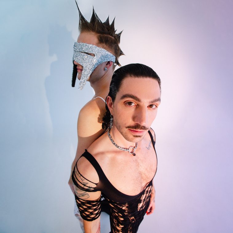 Promotional photo for "Fever Pitch" featuring Conor James that sees BOY2K posing back-to-back, one of the members is wearing a silver visor mask that has two point teeth like a sabretooth tiger, he also has a spiky mohawk. The other member has slicked back black hair, a moustache, and is wearing a black mesh ripped one piece.