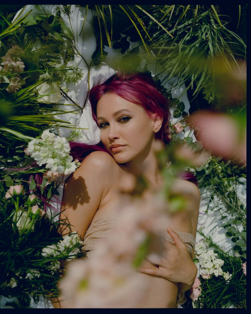 Promotional photo for "holy silence 'fore the spring" EP which sees a head-shot of CLAY lying down amongst grass with various plants in the foreground, as she subtly smiles at the camera. She has purple hair tied back and has a nude-coloured towel over her chest.