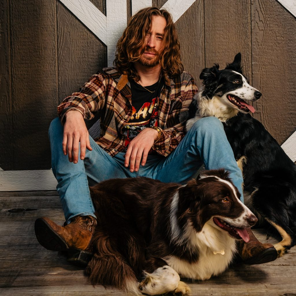 Promotional photo for "Dirt on Me" which sees Colt Graves sitting in front of a stable door, on the ground, wearing a red check shirt over a black tee with blue jeans and cowboy boots. He has shoulder-length dirty blonde hair and a beard. He has two collies around him, a brown collie between his legs, and a black border collie to his left.