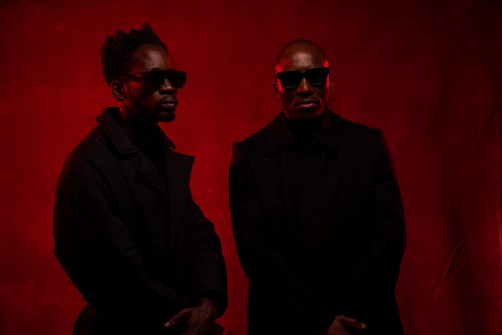 ChopLife SoundSystem posing in front of a red background, side-by-side, wearing black coats and shades.