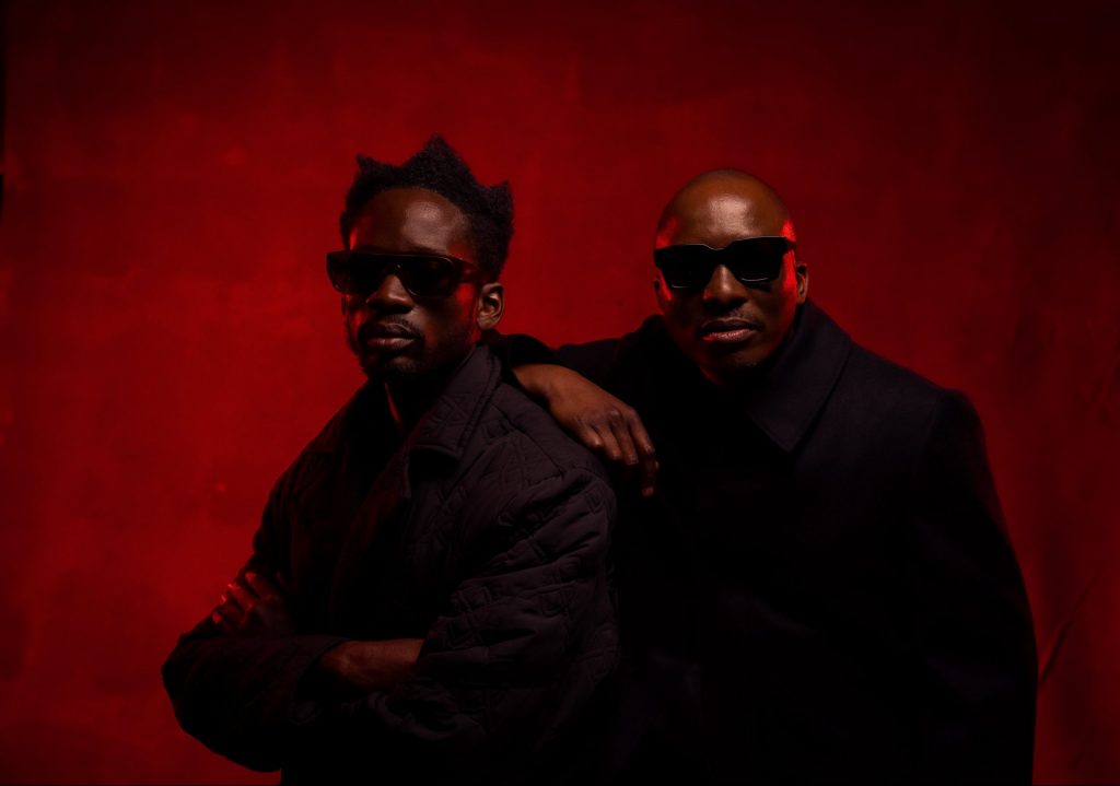 ChopLife SoundSystem posing for the camera in a photoshoot, with DJ Edu leaning his arm across Mr Eazi's back, who has his arms crossed. Both are looking at the camera, wearing black coats, and wearing shades, with a royal red background.