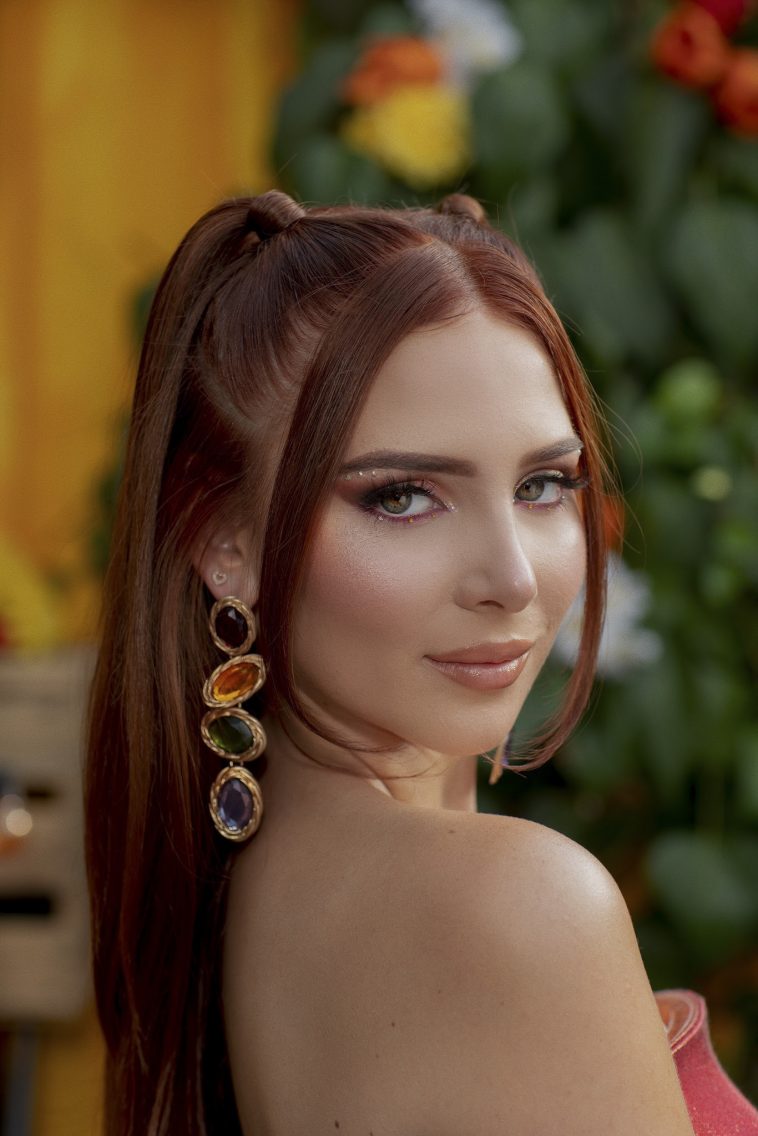 Promotional photo for "No Patience (No Tengo Paciencia)" which sees a head-shot of Chloe Jane looking over her shoulder with a three-gem earring dangling from her ear and her auburn hair tied back into two ponytails with two hair tendrils framing her face.