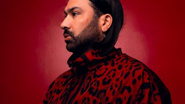 Promotional photo for "Out Of My Mind" featuring Jalja which sees GATTÜSO facing to the left in a headshot with a red background. He has his black hair tucked behind his ear showing off his dark beard, and he is earing a red leopard print jacket that is slightly darker than the background.