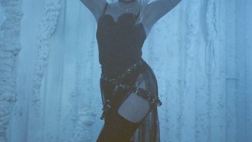 Promotional photo for "King" which sees GG Magree wearing a black silk see-through garment with black boots and a blue wood-like background. She has her hands above her head, just above her blonde mess bob, with her elbows bent.