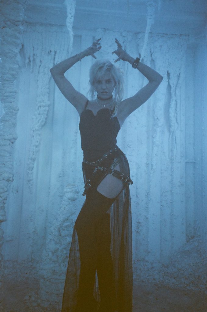 Promotional photo for "King" which sees GG Magree wearing a black silk see-through garment with black boots and a blue wood-like background. She has her hands above her head, just above her blonde mess bob, with her elbows bent.