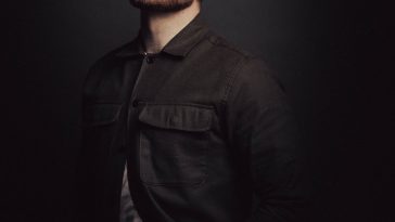 Promotional photo for "All Done" which sees the frontman of GRAYtheband, Gray Ford, posing with his arms behind his back, his eyes closed, and facing towards the left with his chin up. He is wearing a dark shirt over a grey t-shirt, and has a spotlight on him. The background is black and the image is quite dark, highlighting him more.