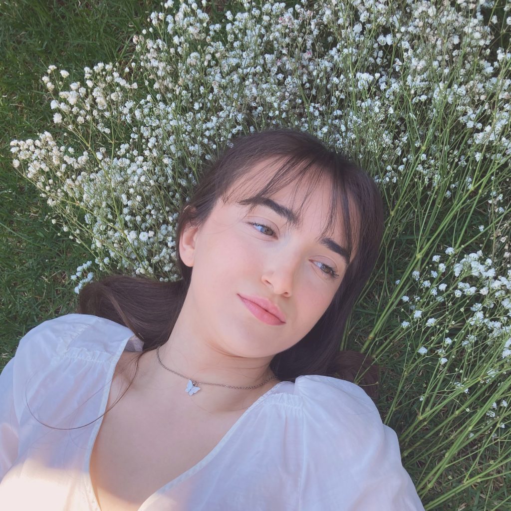 Promotional photo for "Gravity" which sees Jesse Adams lying on a patch of grass with her head brushed against a plot of flowers. She has her brown hair tied back and she's wearing a white open-neck t-shirt.