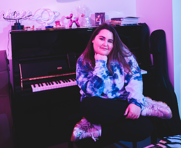 Promotional photo for "Text My Mom" which sees Jules Brave sitting on a piano stool with a black upright piano behind her. The photo has a purple filter to it, and she is wearing a bright tie-dye hoodie with black jeans and white trainers, she is sitting cross-legged.