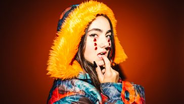 Promotional photo for "All Outta Love" which sees KEHLI standing in front of a dark-orange backdrop, wearing a multi-coloured puff coat with the hood up on her head, which has a bright orange faux-fur look to it. Her make-up is pure white with three upside-down red heart gems placed in a vertical line down her eyes and over her cheeks.