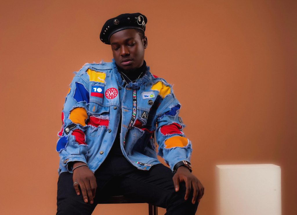 Press photo for "Shy" which sees Killertunes in a blue denim jacket filled with various primary colour shades of patches, an artist's black cap with matching trousers and shirt, in promotion of the collaboration with BZ featuring Not3s.