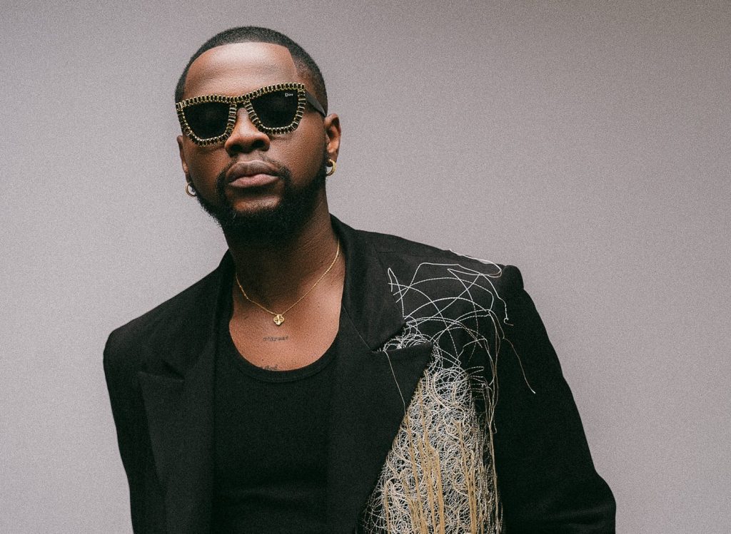 Promotional photo for "Shu-Peru" which sees Kizz Daniel wearing a black smart jacket with fashion string attached to the right side, over a black vest top. He is wearing some gold-outlined shades and he is standing in front of a grey background.