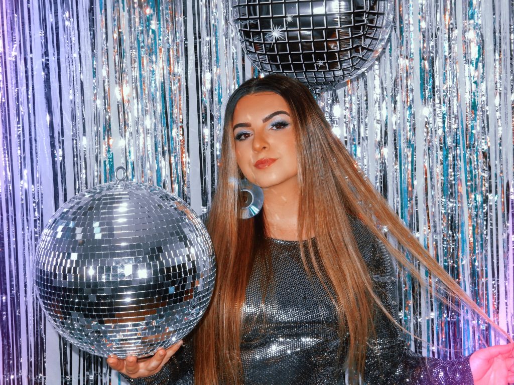 Promotional photo for "No Worries..." which sees Lexi Mariah surrounded by mirror balls and a silver shimmery curtain behind her. She's wearing a silver dress and her blonde-brown hair flows down past the photo. She's wearing CD earrings, and she's holding a mirror ball in her hand.