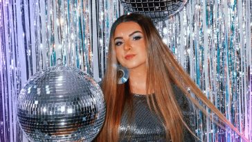Promotional photo for "No Worries..." which sees Lexi Mariah surrounded by mirror balls and a silver shimmery curtain behind her. She's wearing a silver dress and her blonde-brown hair flows down past the photo. She's wearing CD earrings, and she's holding a mirror ball in her hand.