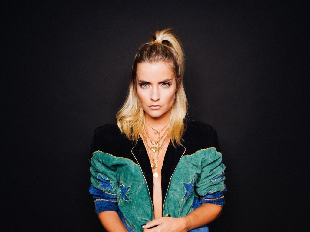 Promotional professional headshot photograph for "The House of House" which sees LOVRA standing in front of a black background, with her blonde hair tied up in a half-ponytail with the rest of her hair falling just past her shoulders and around her blue and black zipped jacket that is unzipped and revealing her flesh.