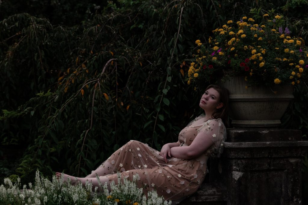 Promotional photo for "Castles" which sees PiPEllA relaxing in a garden with her back leaning against a tree. She is earing a light-pink floor-length dress.