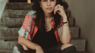 Promotional photo for "Open Wide" which sees Tatiana DeMaria sitting on some concrete steps with her dark hair ruffled, which flows over her salmon and green shirt which is unbuttoned over a black tank top. She pairs all that with faded-black ripped jeans. She has her left elbow on her left leg and she's resting her head slightly on her left hand, whilst the right arm is just resting on her right leg.