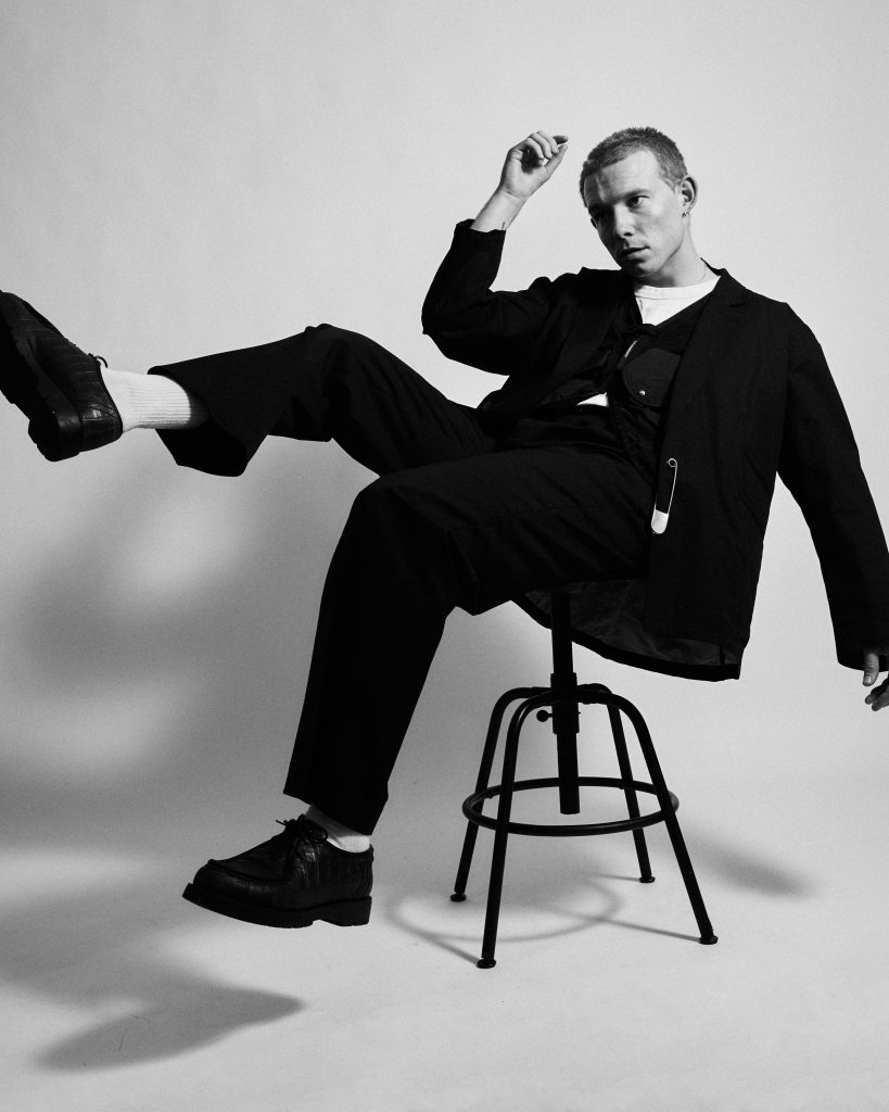 A black and white promotional photo for "Colours" that sees VC Pines chilling on a stool with a tilted appearance with one leg in the air and his hand up but bent at the elbow. He is dressed smartly and is exposing his beautiful jawline by looking to the far-left.
