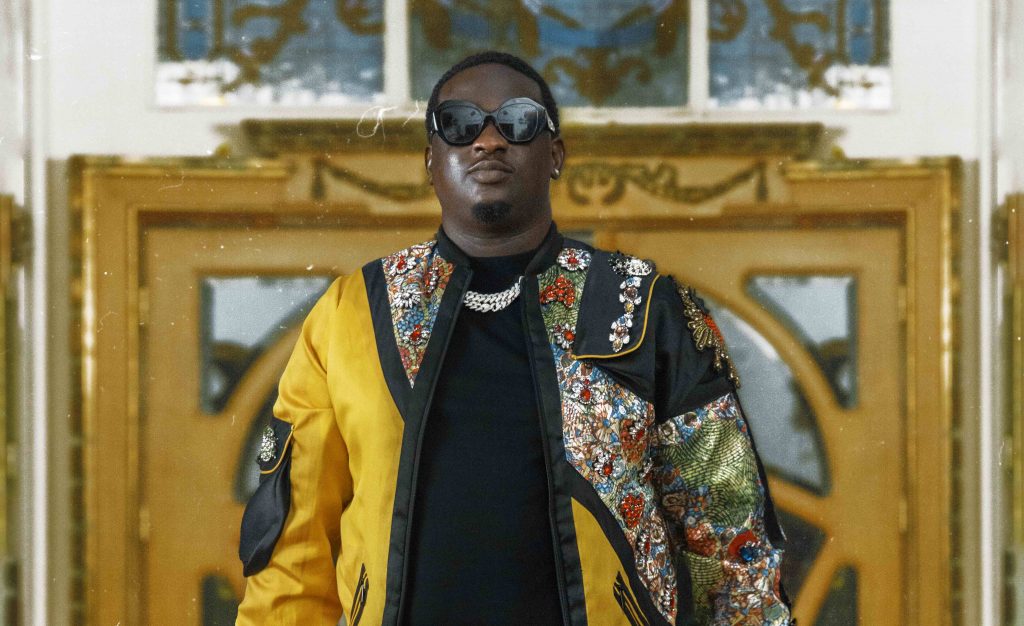 Promotional photo for "Let Them Know" which sees Wande Coal wearing a black t-shirt under a gold chain necklace and a yellow jacket that has African prints cascading from the top left to the bottom right of the jacket. He is wearing black shades, and is standing in front of a ornate wooden door.