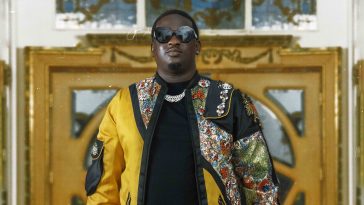 Promotional photo for "Let Them Know" which sees Wande Coal wearing a black t-shirt under a gold chain necklace and a yellow jacket that has African prints cascading from the top left to the bottom right of the jacket. He is wearing black shades, and is standing in front of a ornate wooden door.
