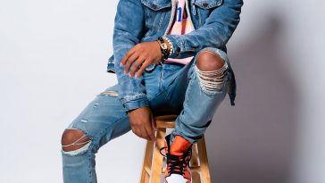 Promotional photo for "She Wants To Love" which sees Adam Ford sitting on a wooden stool wearing double denim with ripped jeans and a denim jacket over a pink hoodie. He's also wearing orange and white Nikes.