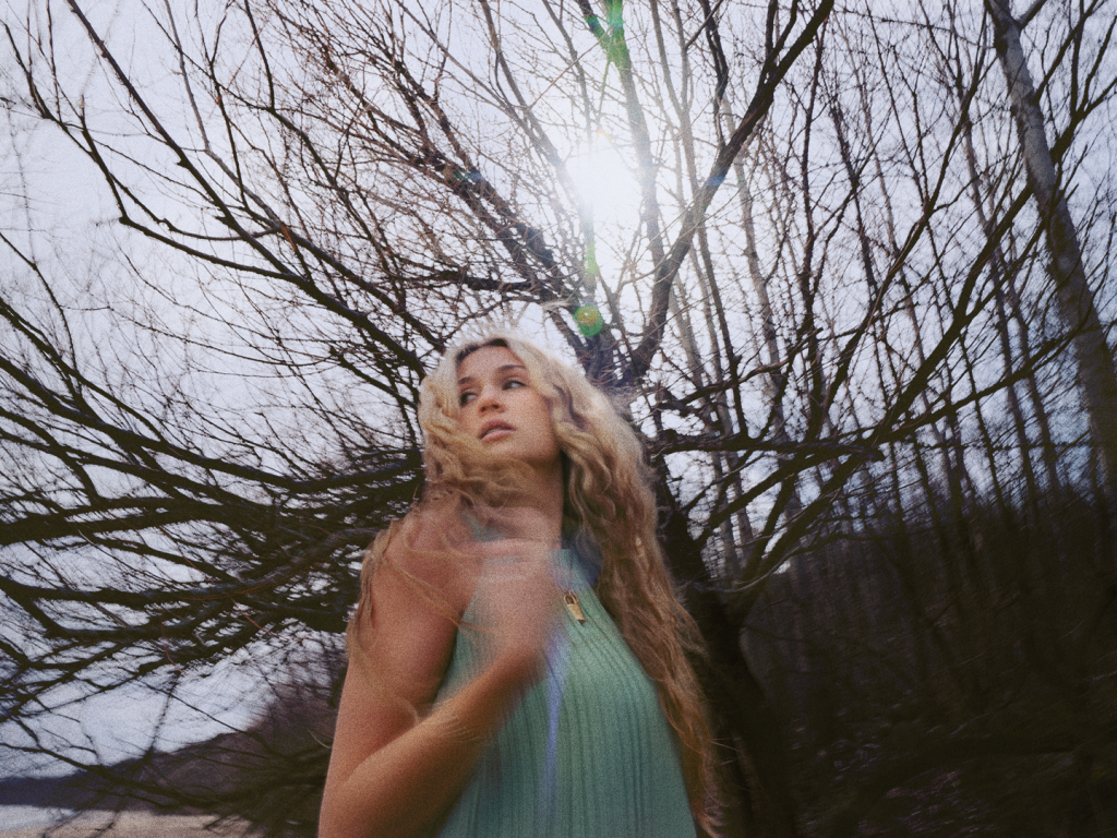 Promotional photo for "Heavy, Heavy, Heavy" which is a mid-shot of Ashley Elle in the woods with the camera tilted upwards. She's wearing a teal dress and the camera has caught her moving her head so her cascading blonde hair is moving, blurred in the shot, making her look whimsical.