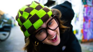 Promotional photo for "Get This" which sees Belot leaning forward towards the camera, in the street, wearing a green and black checkered beanie, black-framed glasses, and a black t-shirt. She is grinning.