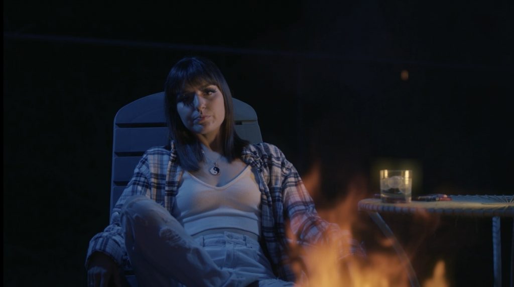 Still from "Her and I" music video which sees Calliope Wren sitting on a wooden backyard chair with flames in front of her and a wooden table to the right of her. She is wearing an unbuttoned checkered cream and blue shirt, a white vest top, blue ripped jeans, and a necklace with the yin and yang peace symbol.