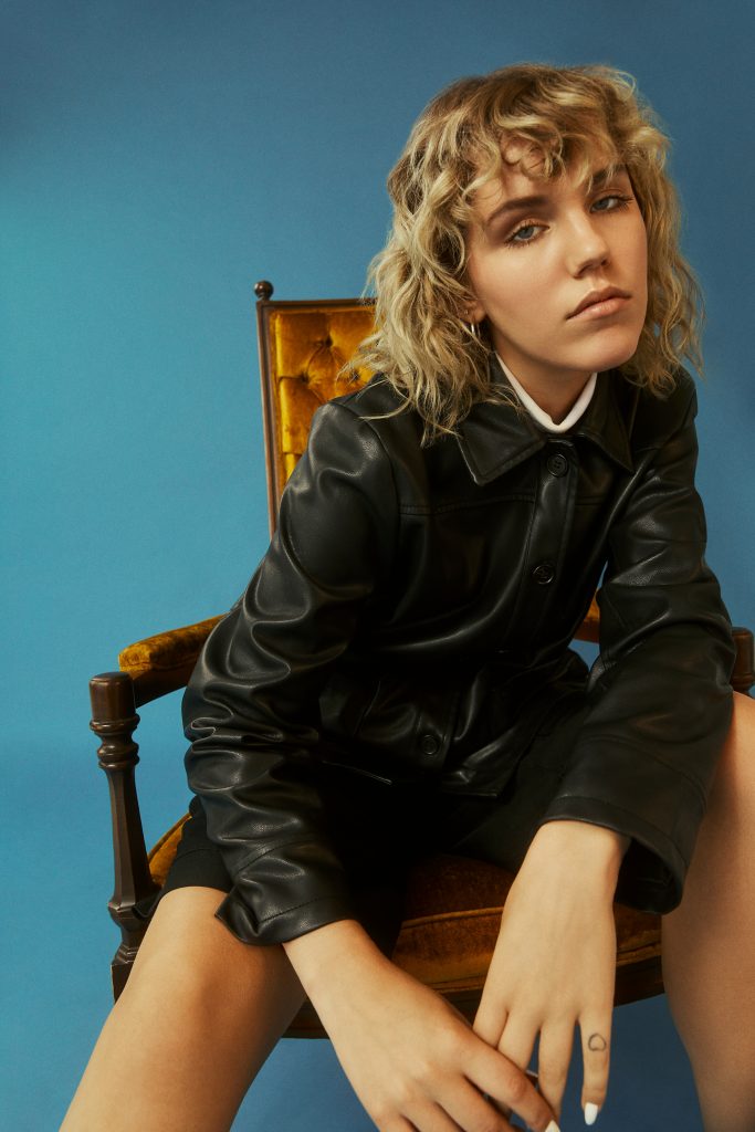 Promotional photo for "Try To Get Away" which sees Carlyn sitting on a wooden chair in front of a blue wall, wearing a black leather jacket, over some black shorts. Her blonde hair falls as waves to her shoulders.