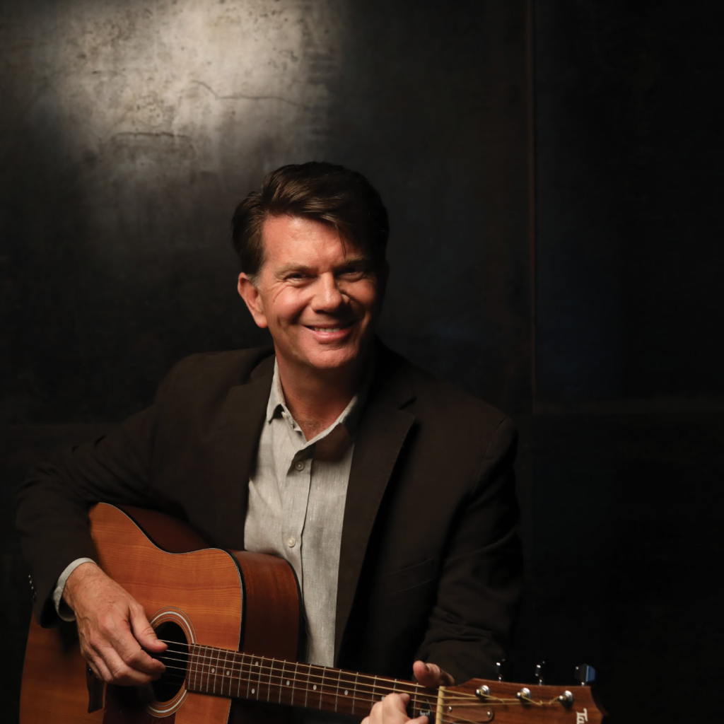 Promotional image for "The Sinner and the Saint" which sees Chris St John sitting, playing a guitar, in front of a black background. He is wearing a white shirt under a black suit jacket.