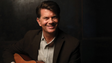 Promotional image for "The Sinner and the Saint" which sees Chris St John sitting, playing a guitar, in front of a black background. He is wearing a white shirt under a black suit jacket.