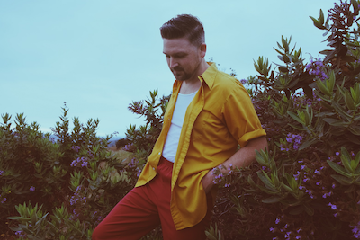 Promotional photo for "Lost In The Rhythm" which sees Daniel Ellsworth wearing a white vest under an open mustard-yellow shirt and burnt-orange trousers, leaning backwards in a bush.