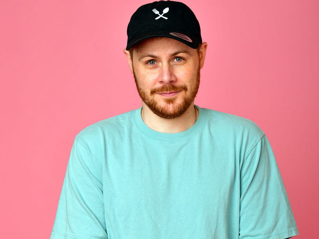 Headshot image of Spag Heddy wearing a baby blue sweater that contrasts perfectly with the baby pink background. He is also wearing a black snapback cap and he is smiling.
