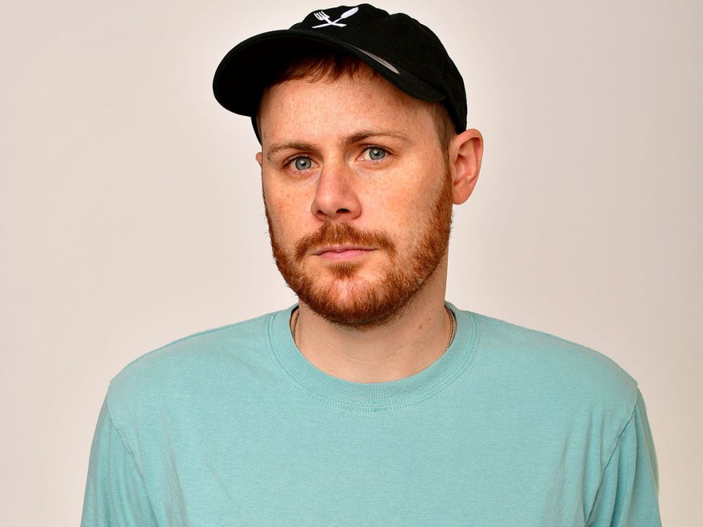 Headshot image of Spag Heddy wearing a baby blue sweater with a black snapback cap. The background is a light grey, and his head is slightly tilted to the left.
